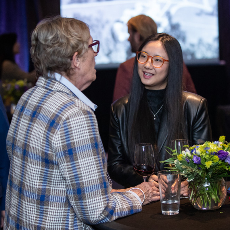 Two people standing at a cocktail table during a reception, in an event space with a screen in the background.